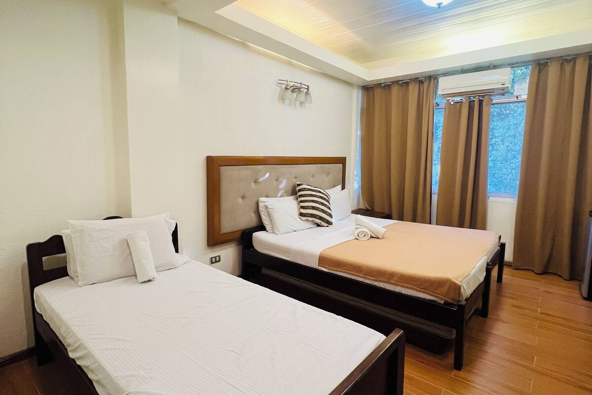 A hotel room with two beds, a ceiling light, an air conditioning unit, and a window with curtains.