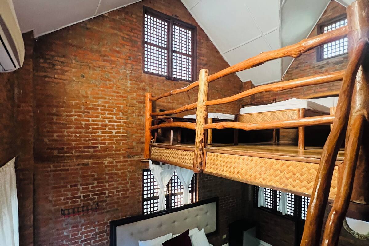 A rustic room with a brick wall, a wooden bunk bed, a single bed with white bedding, and a small table.