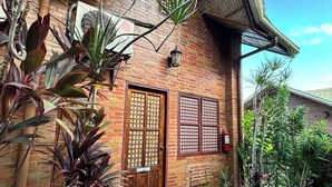 Un bâtiment traditionnel en briques, doté d’une porte en bois et de volets aux fenêtres, entouré d’une végétation tropicale luxuriante.