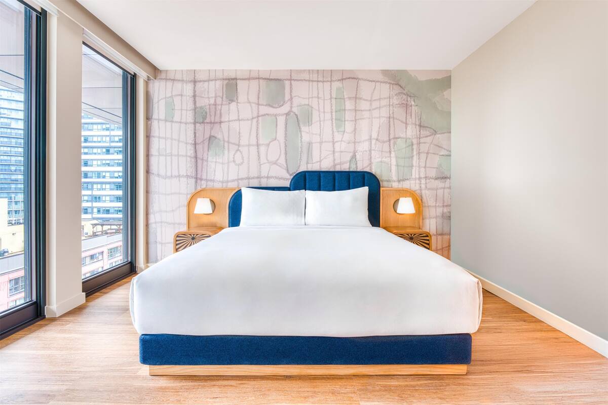 Ein modernes Hotelzimmer mit einem großen Bett, einem Holzkopfstück und Blick auf die Stadt durch große Fenster.