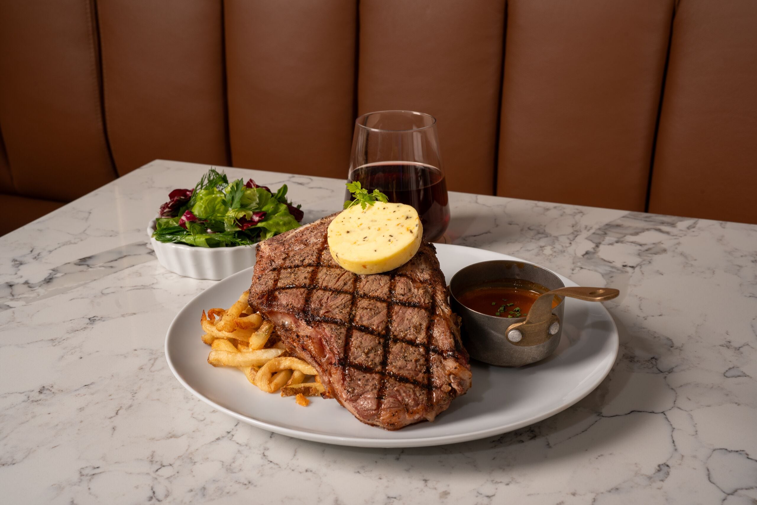 A plate with a grilled steak, fries, and a salad, accompanied by a glass of red wine and a small bowl of sauce.