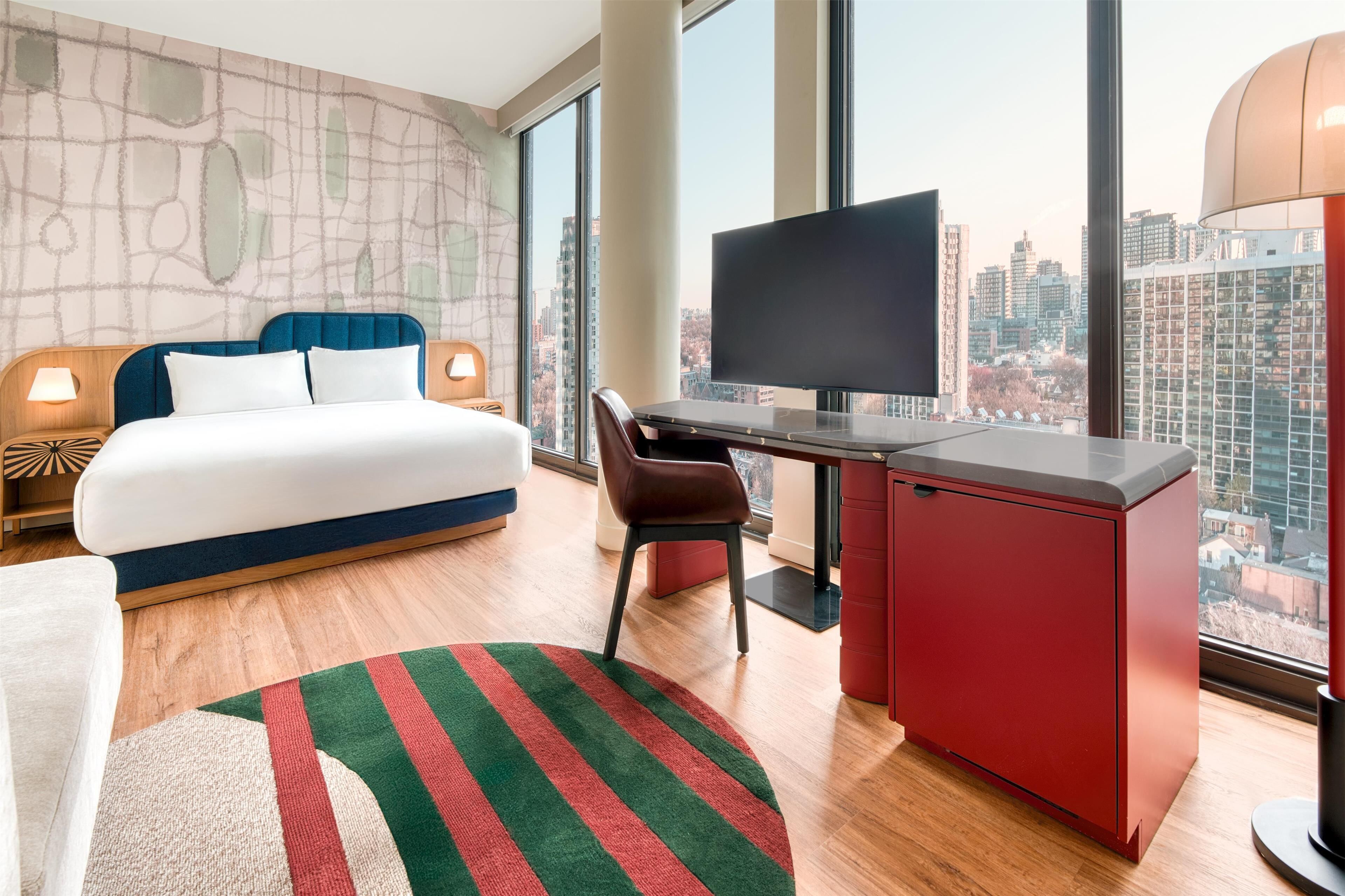 A hotel room with a bed, desk, chair, TV, and large windows with a city view.