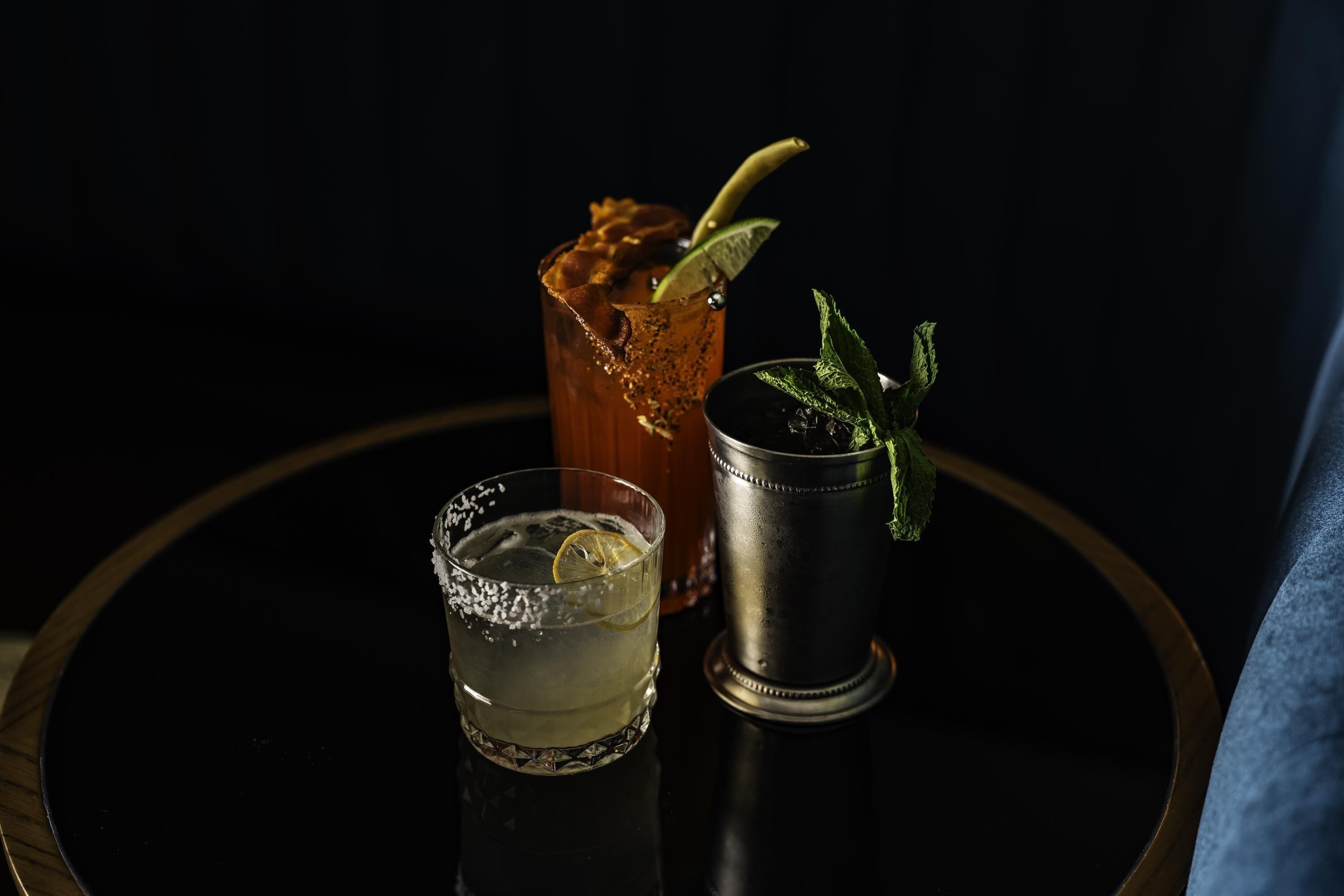 A round table with three drinks: a cocktail glass, a highball glass, and a metal cup.
