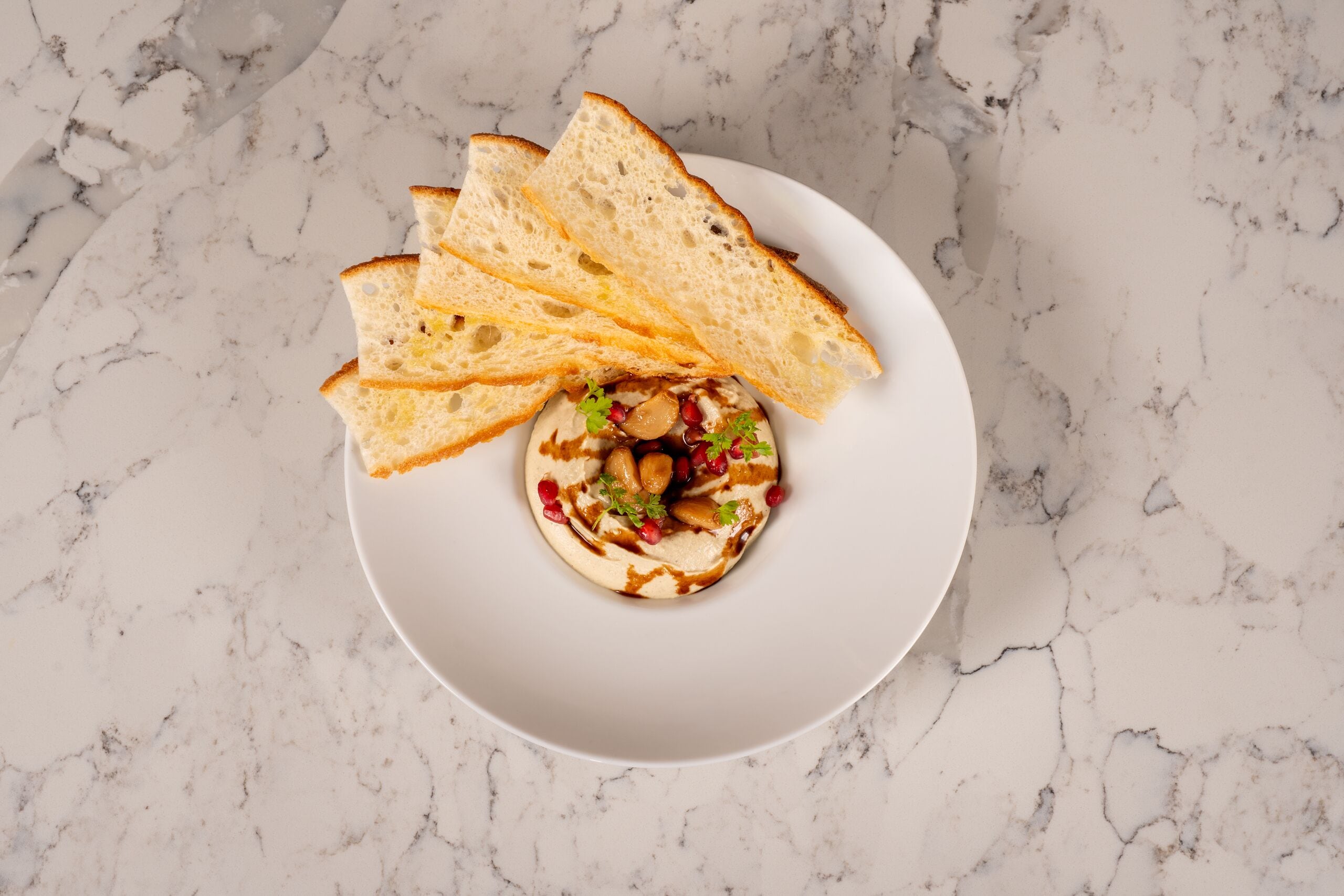 A white plate with a dollop of hummus and pomegranate seeds on a marble surface.