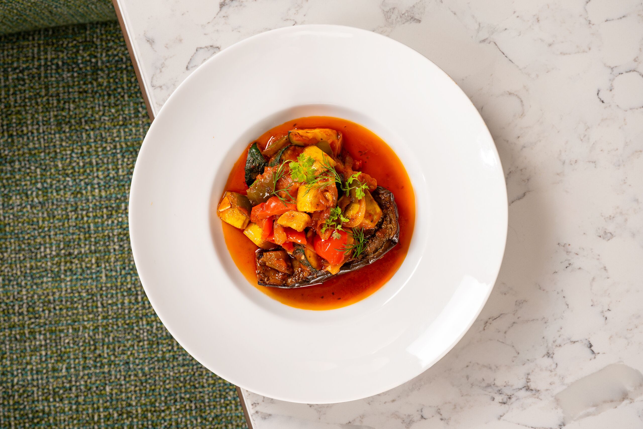 A white plate with a dish of vegetables and a tomato-based sauce on a marble countertop.