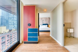 Een moderne hotelkamer met een groot raam, een kast in rood en blauw, een bed met blauw-witte beddengoed en een metalen handdoekrek.