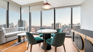 Ein modernes Wohnzimmer mit einem runden Tisch, grünen Stühlen, einem grauen Sofa und großen Fenstern mit Blick auf die Stadt.