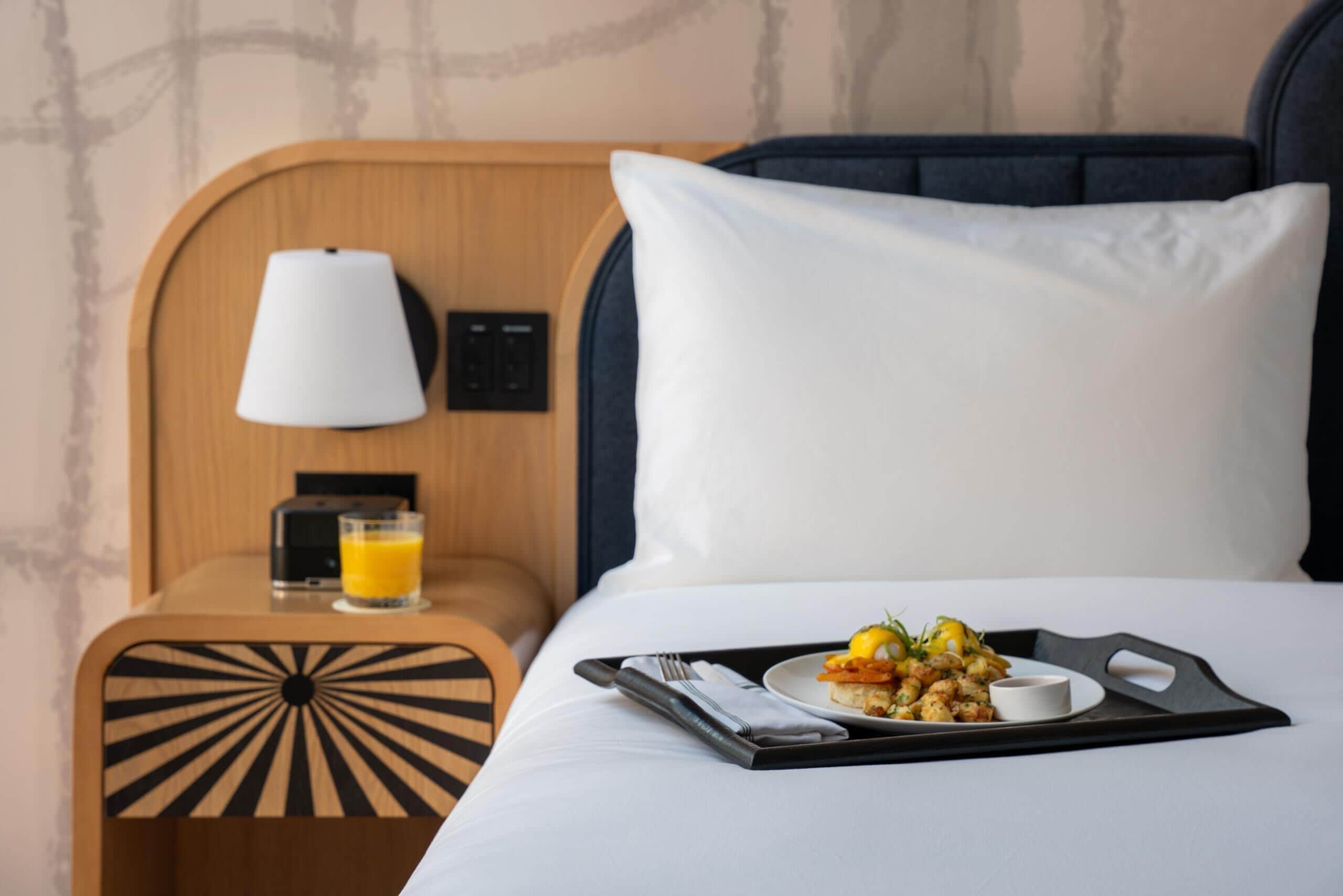 A neatly made bed with a tray of food and a glass of orange juice on a tray, a bedside table with a lamp, and a power outlet.