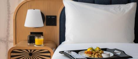 A neatly made bed with a tray of food and a glass of orange juice on a tray, a bedside table with a lamp, and a power outlet.