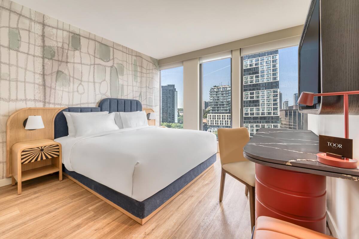 Ein modernes Hotelzimmer mit einem großen Bett, einem Nachttisch aus Holz, einem roten Akzentstuhl, einem Tisch mit Marmorplatte und Blick auf die Stadt durch große Fenster.