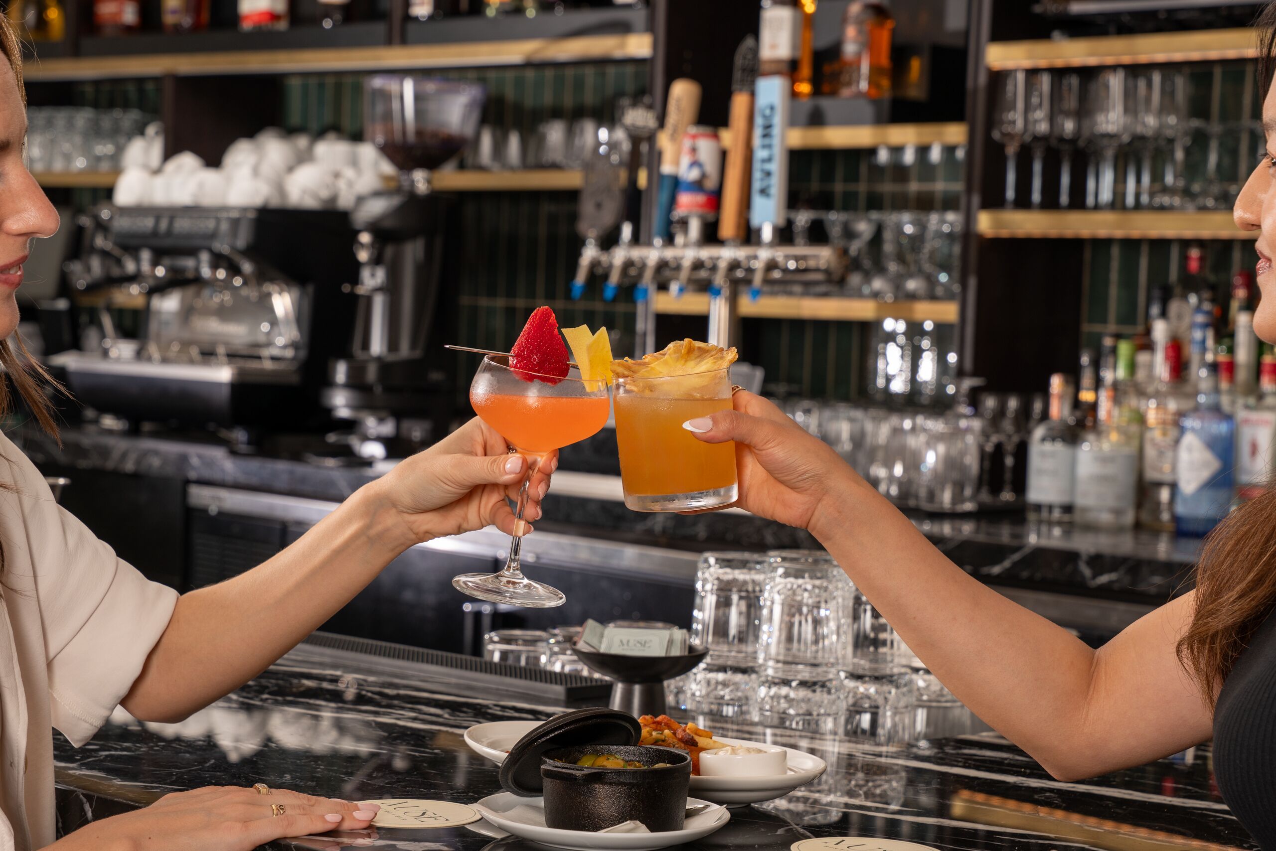 Un bar con dos personas brindando con bebidas, una máquina de café y una variedad de botellas al fondo.