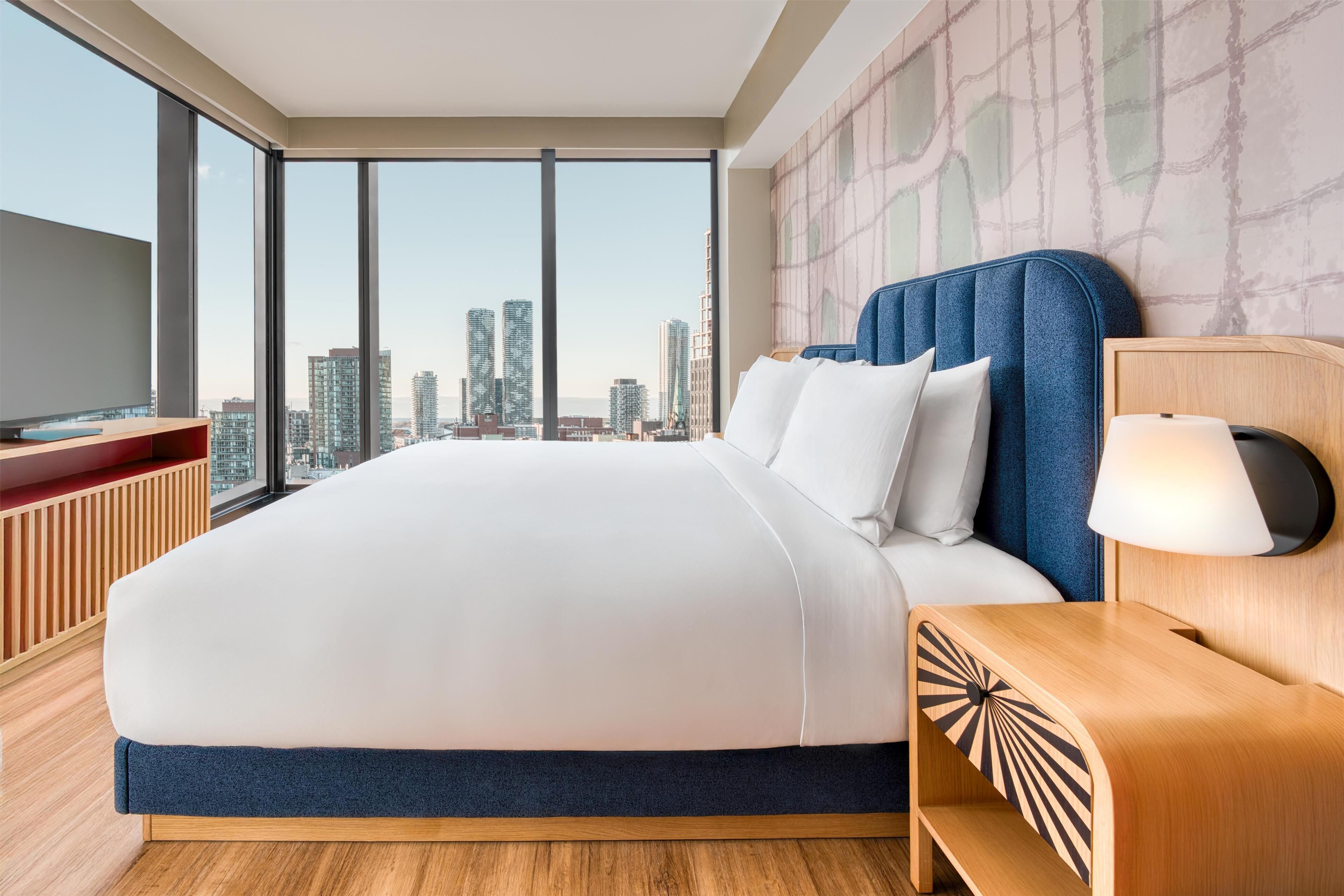 Una habitación de hotel moderna con una cama grande, vista a la ciudad y una mesita de noche de madera.