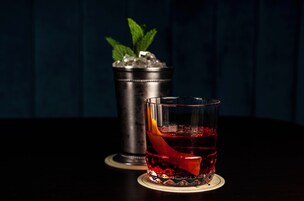 Een cocktailglas met een rode drank en een oranjeschijfje op een onderzetter, een metalen tumbler met ijs en muntblaadjes, en een donkerblauwe achtergrond.
