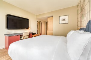 Habitación de hotel con una cama grande, un escritorio con silla, un televisor de pantalla plana, un cuadro en la pared y una puerta de madera.