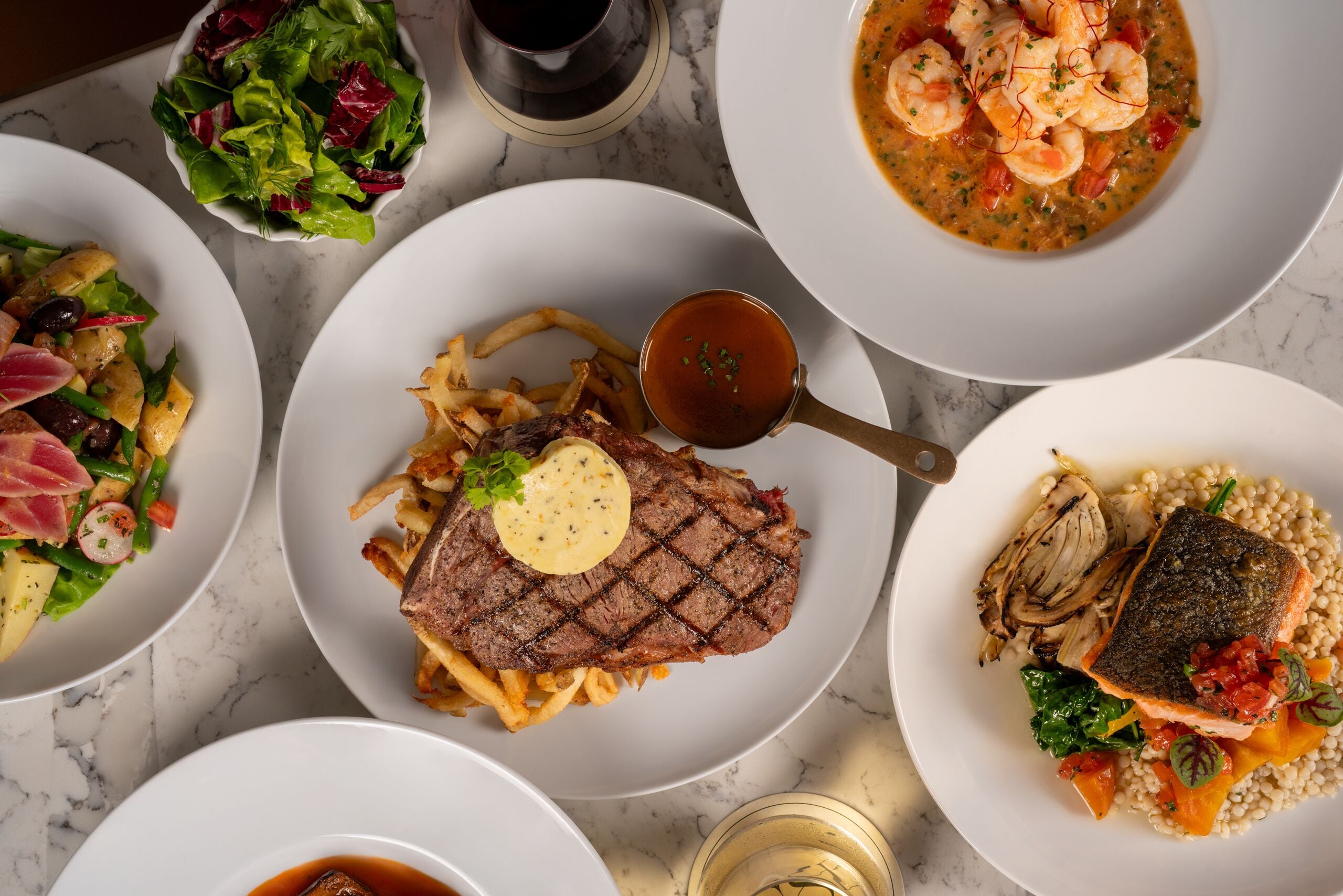 A variety of dishes on a marble table, including steak, fish, and a shrimp soup.