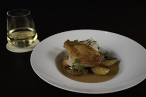 Een wit bord met vlees, geroosterde aardappelen en een takje peterselie, vergezeld van een glas witte wijn op een donkere achtergrond.