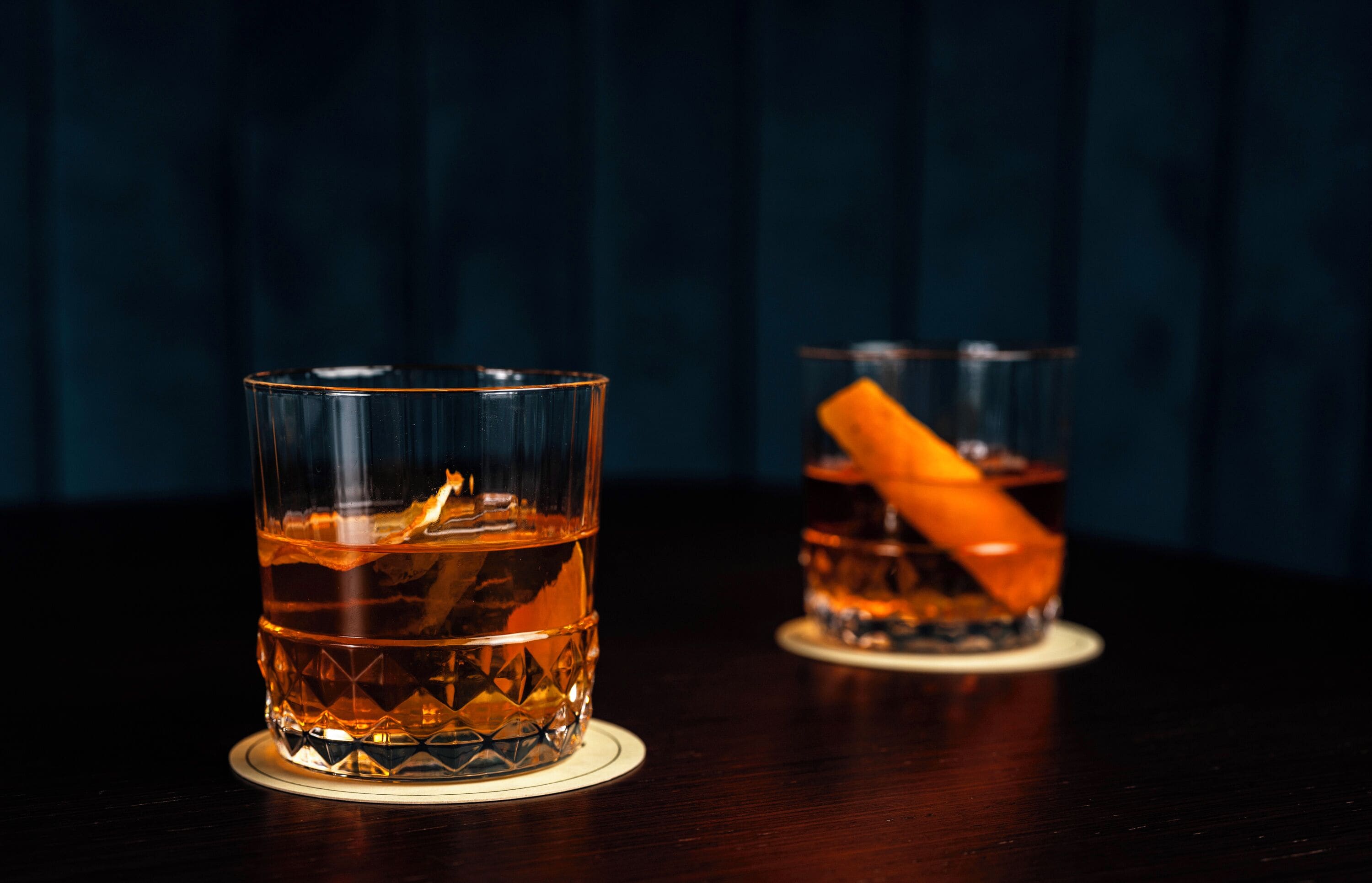 Two glasses of amber liquid on a dark wooden table with a blue backdrop.