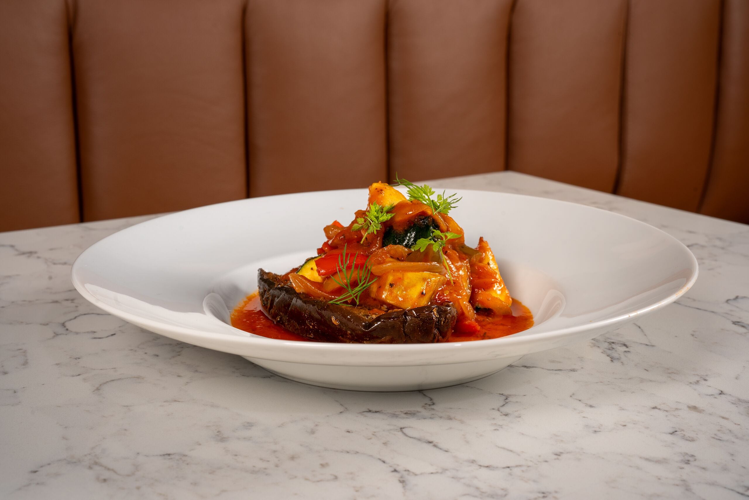 Une assiette blanche sur laquelle se trouve un plat d’aubergines farcies, le tout posé sur une table en marbre.