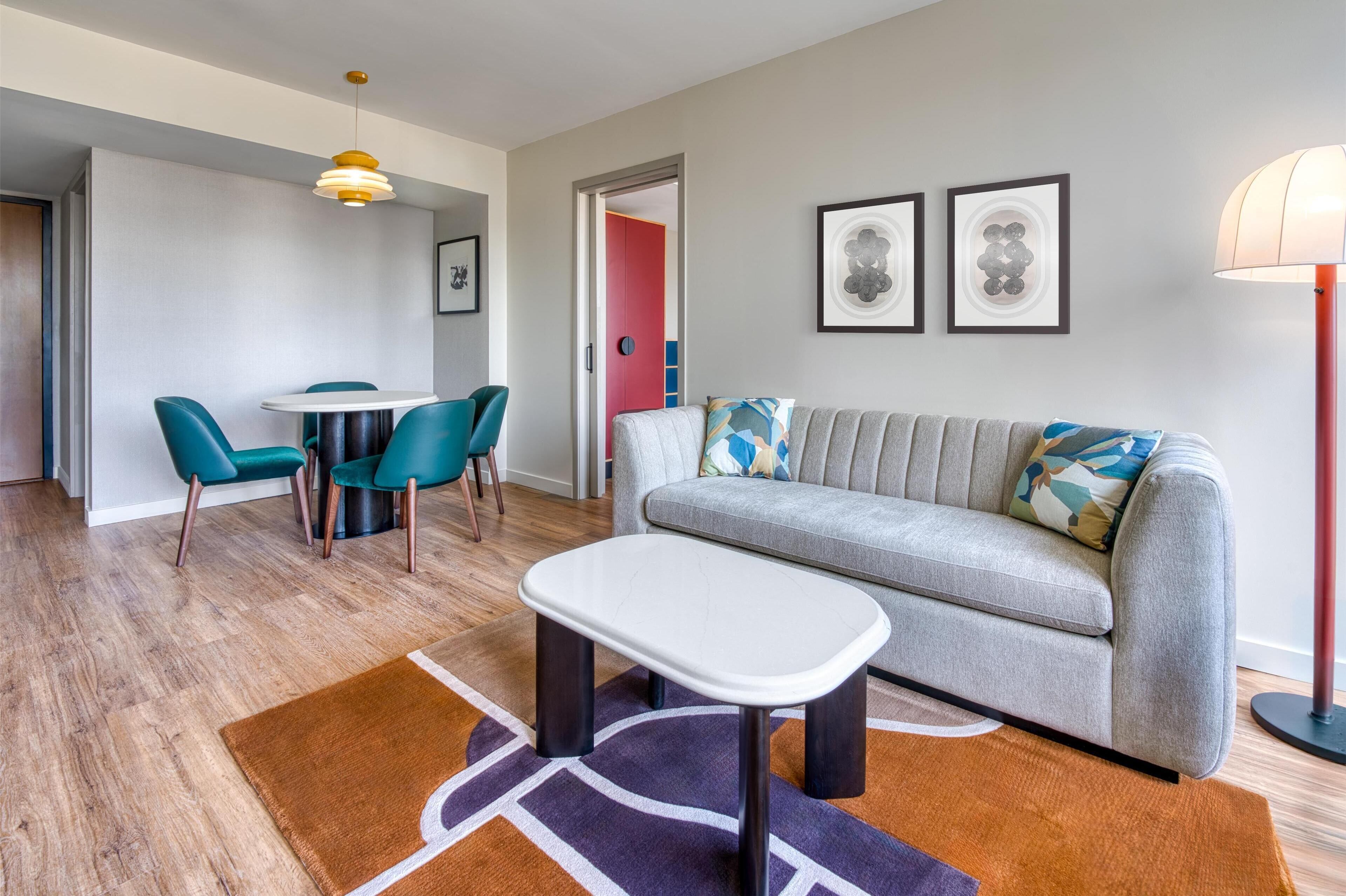 Ein modernes Wohnzimmer mit einem grauen Sofa, smaragdgrünen Sesseln, einem weißen Couchtisch und einem gemusterten Teppich.