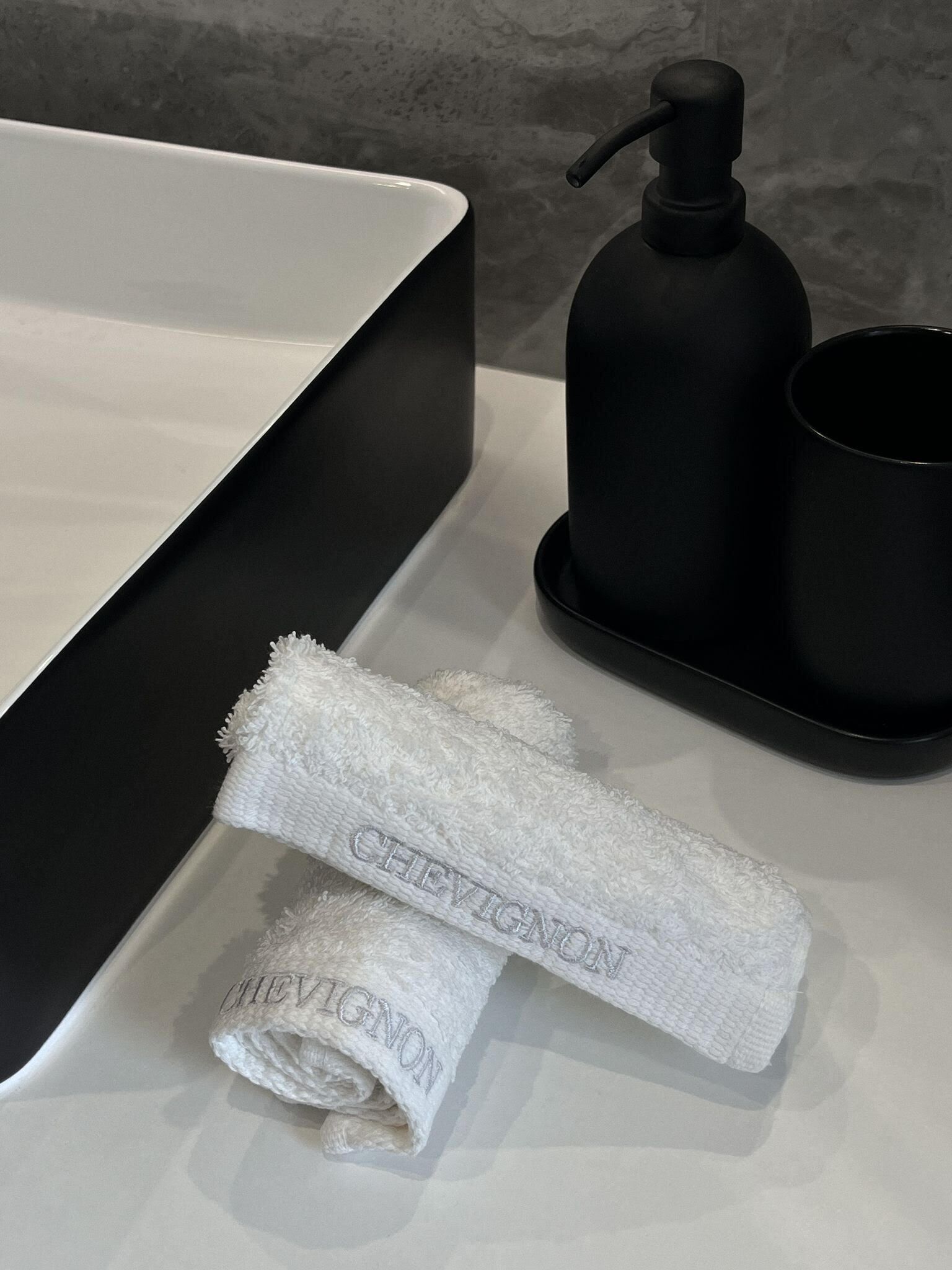 Un baño con lavamanos blanco, dispensador de jabón negro y dos toallas blancas con el bordado 'CHEVIGNON'.