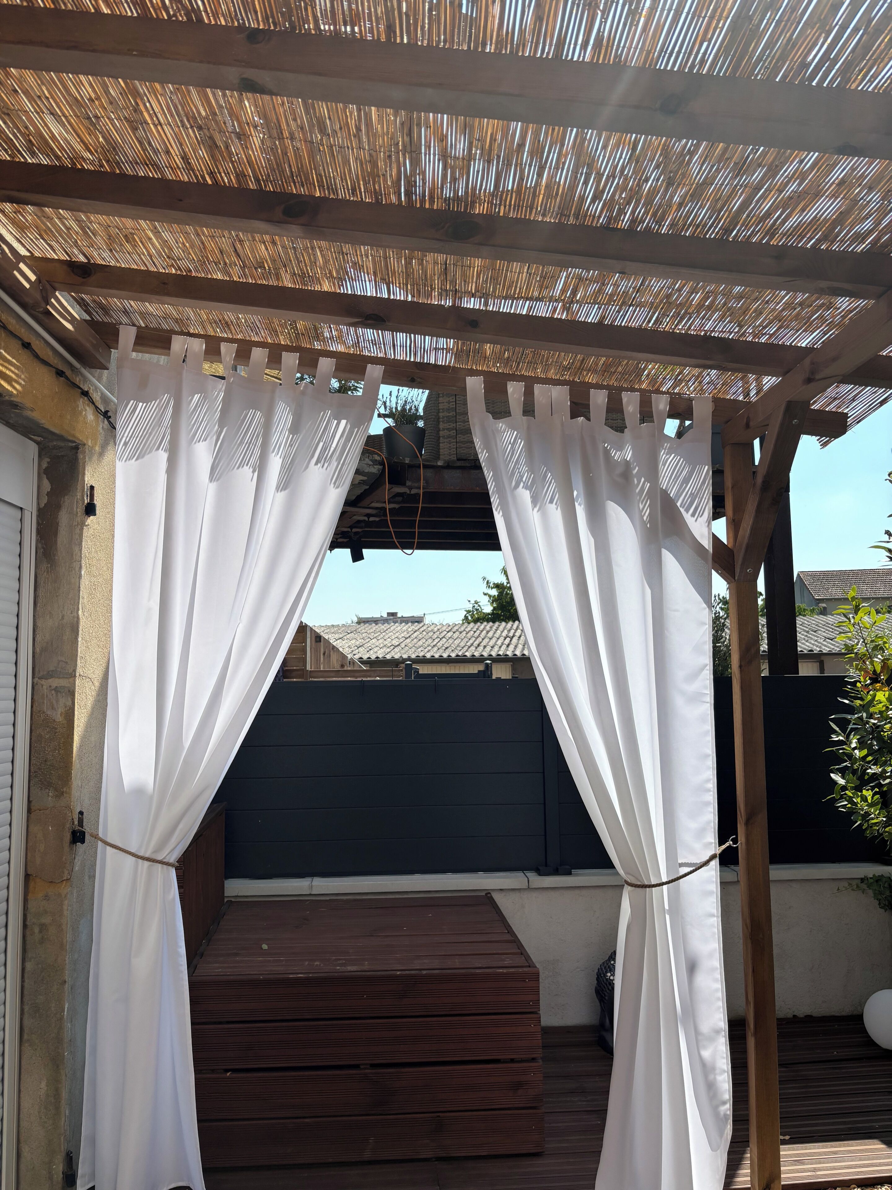 Un espace extérieur abrité doté d’une terrasse en bois, de rideaux blancs et d’une clôture noire.