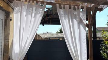 Un espace extérieur abrité doté d’une terrasse en bois, de rideaux blancs et d’une clôture noire.