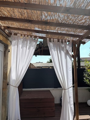 A covered outdoor area with a wooden deck, white curtains, and a black fence. - Arum Suite with Terrace (Roanne)