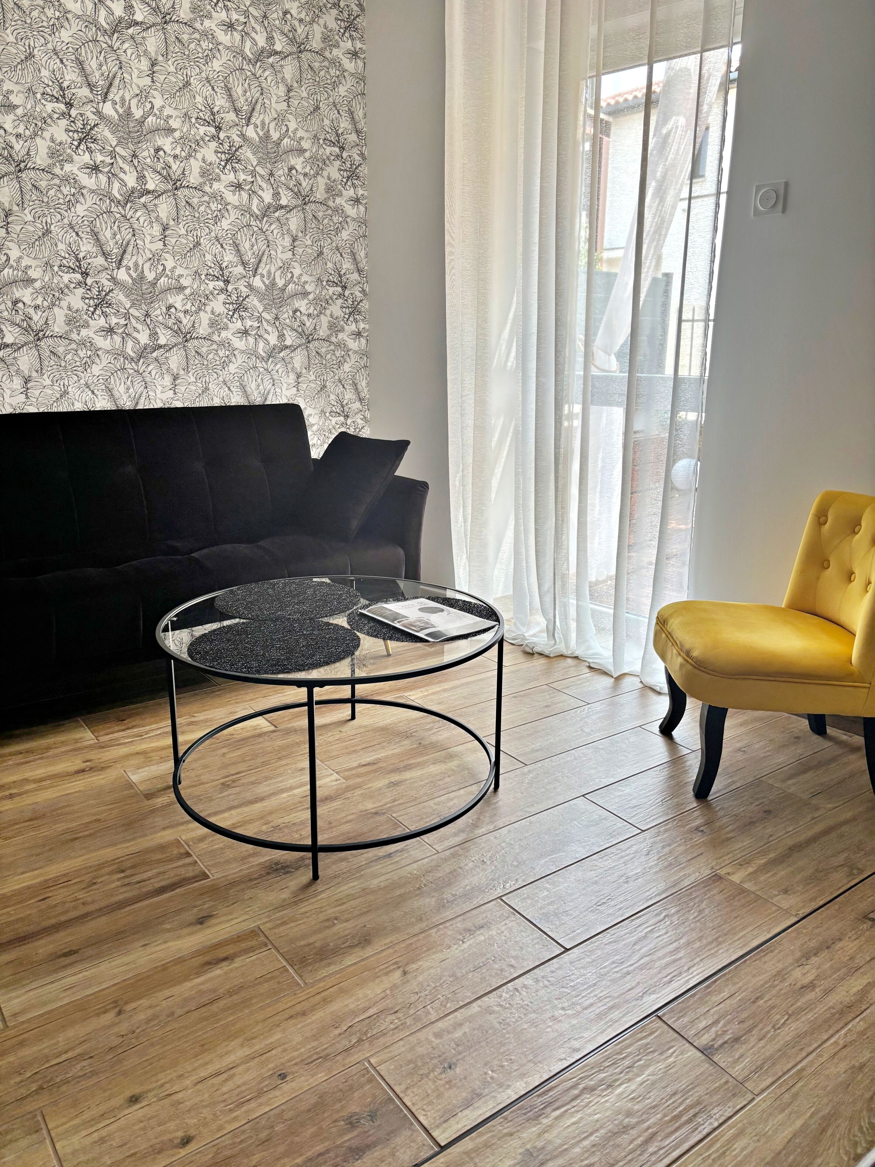 Een woonkamer met een zwarte bank, een gele fauteuil, een glazen salontafel en behangpapier met een patroon.