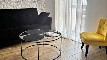 Een woonkamer met een zwarte bank, een gele fauteuil, een glazen salontafel en behangpapier met een patroon.