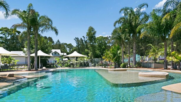 A clear swimming pool surrounded by palm trees and white umbrellas, with a fence and a building in the background. - Wild Duck Holiday Home at Rafferty's Resort (Cams Wharf)