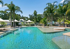 A clear swimming pool surrounded by palm trees and white umbrellas, with a fence and a building in the background. - Wild Duck Holiday Home at Rafferty's Resort (Cams Wharf)