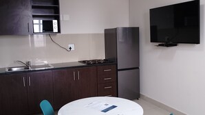 Cocina moderna con alacenas de madera oscura, microondas, estufa, refrigerador y televisor de pantalla plana montado en la pared. Hay una mesa redonda con dos sillas color turquesa y una encimera blanca.