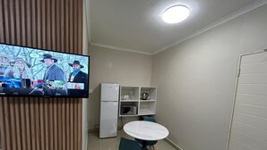 Una cocina pequeña y funcional con un refrigerador blanco, un microondas y un armario blanco. Hay una mesa redonda con dos sillas y un televisor montado en la pared.