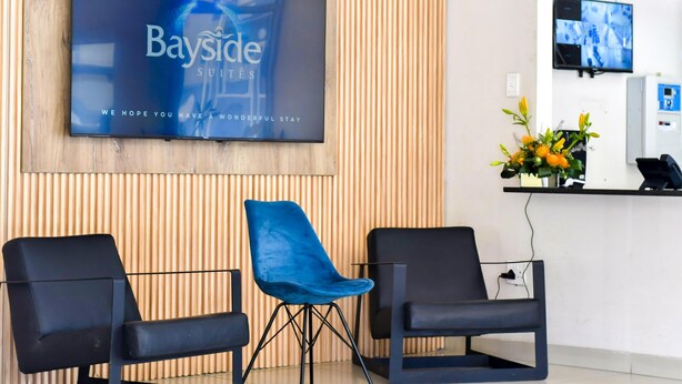 Un área de recepción con el letrero de Bayside Suites, dos sillas negras, una pared de madera y una pantalla de televisión.