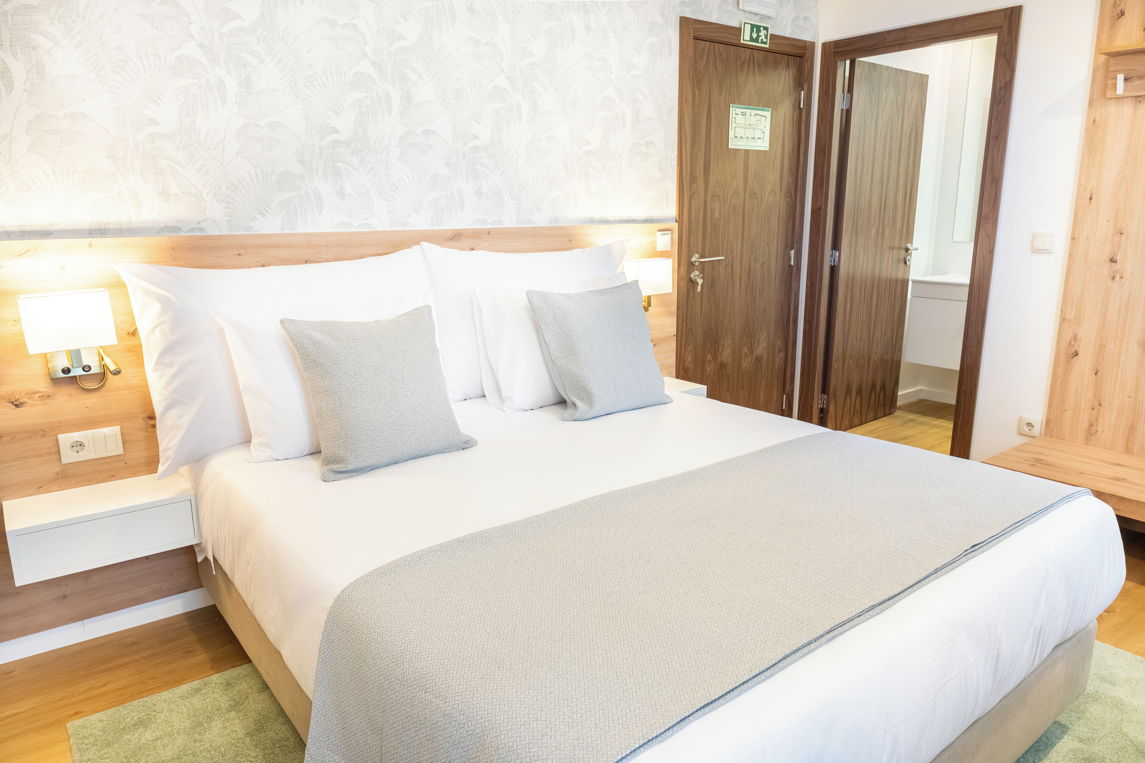 Una cama bien hecha con ropa de cama blanca y gris, un cabecero de madera y una alfombra verde. Hay una puerta de madera con un letrero y una mesita de noche de madera con una lámpara.