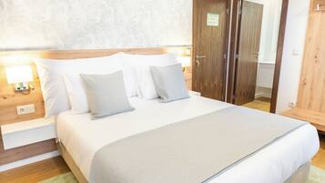 Una cama bien hecha con ropa de cama blanca y gris, un cabecero de madera y una alfombra verde. Hay una puerta de madera con un letrero y una mesita de noche de madera con una lámpara.