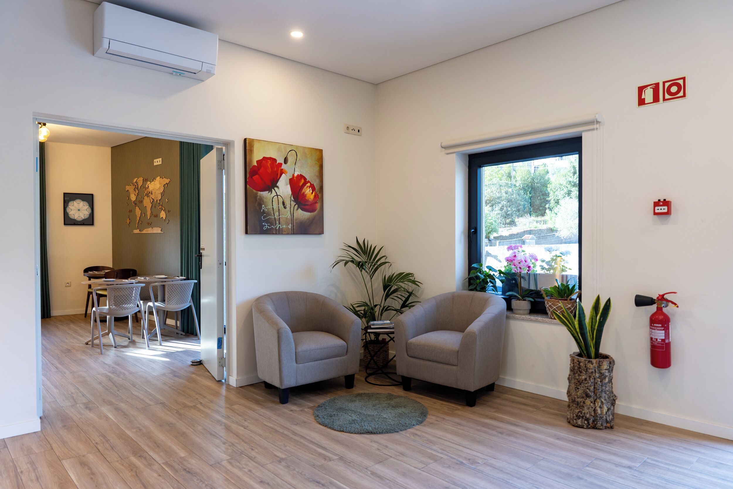 Uma sala de estar moderna com dois poltronas, uma área de jantar com mesa redonda, e uma janela grande com plantas.
