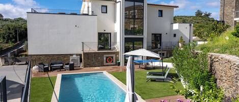 Una casa moderna de dos pisos con piscina, mobiliario exterior y un muro de piedra.