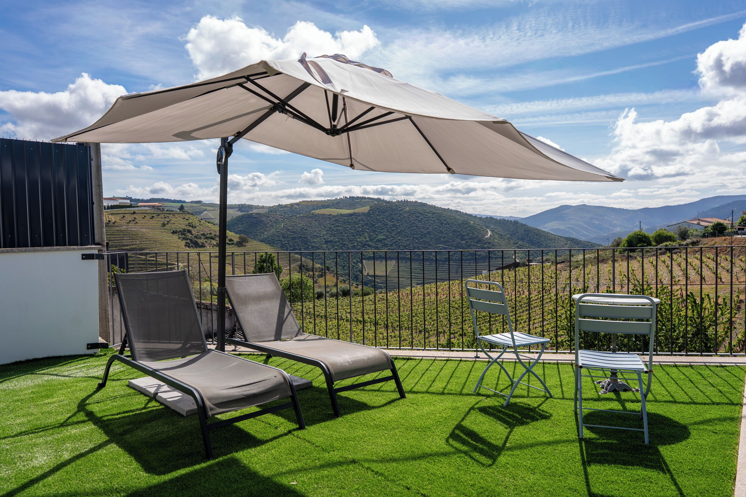 A patio with two lounge chairs, a table, and an umbrella, overlooking a vineyard and hills.