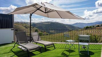 A patio with two lounge chairs, a table, and an umbrella, overlooking a vineyard and hills.