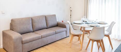 A modern living room with a grey sofa, a dining table with chairs, and a vase with dried plants.