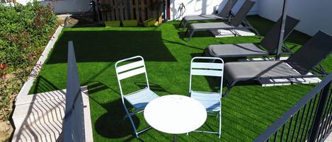 Un patio con césped artificial, dos sillas, una mesita y un paraguas blanco.