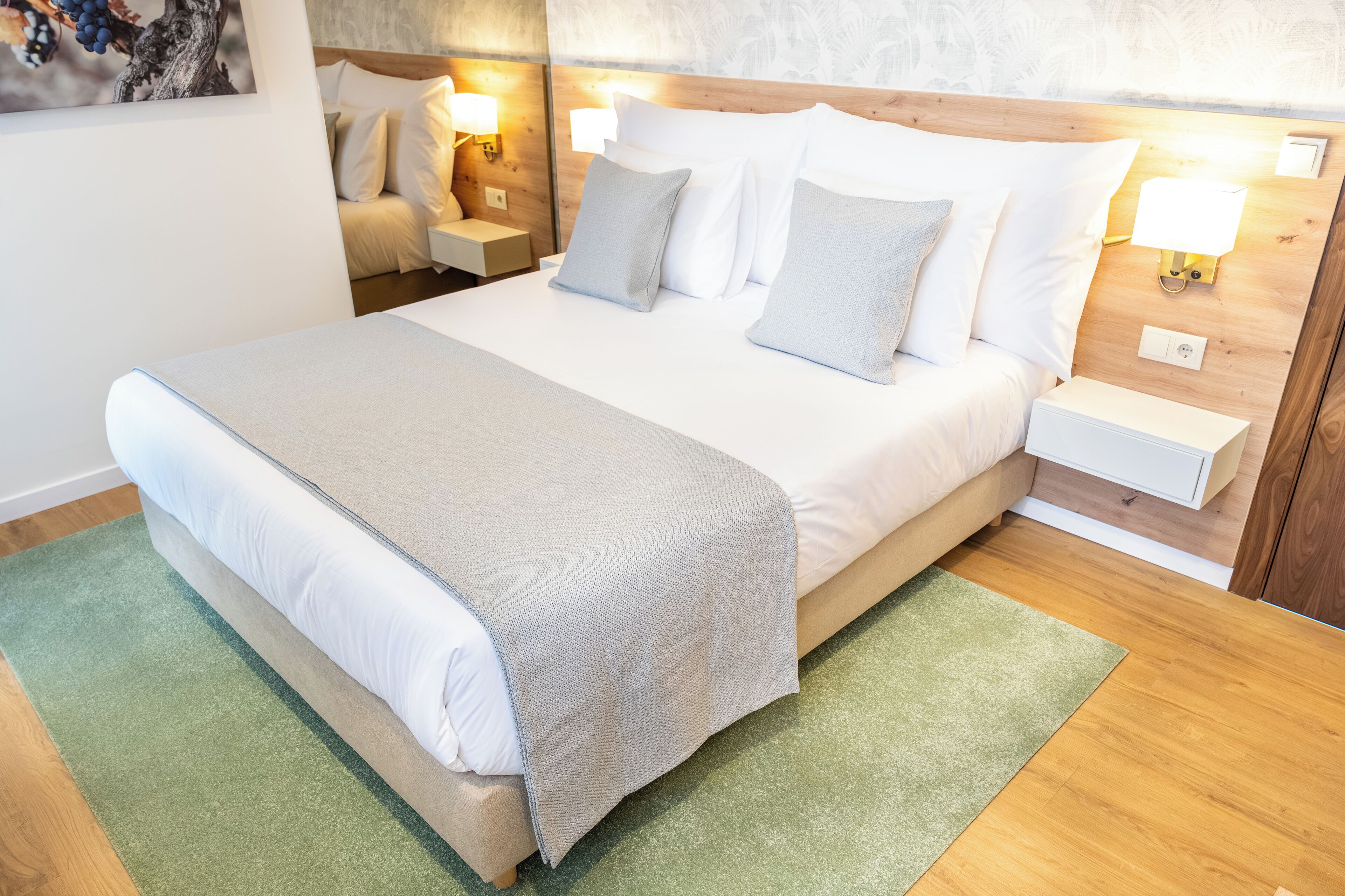Ein ordentlich bezogenes Bett mit heller Bettdecke und Kissen, ein grüner Teppich, ein Holz-Kopfteil, Nachttischlampen und ein Nachttisch mit Lampe.
