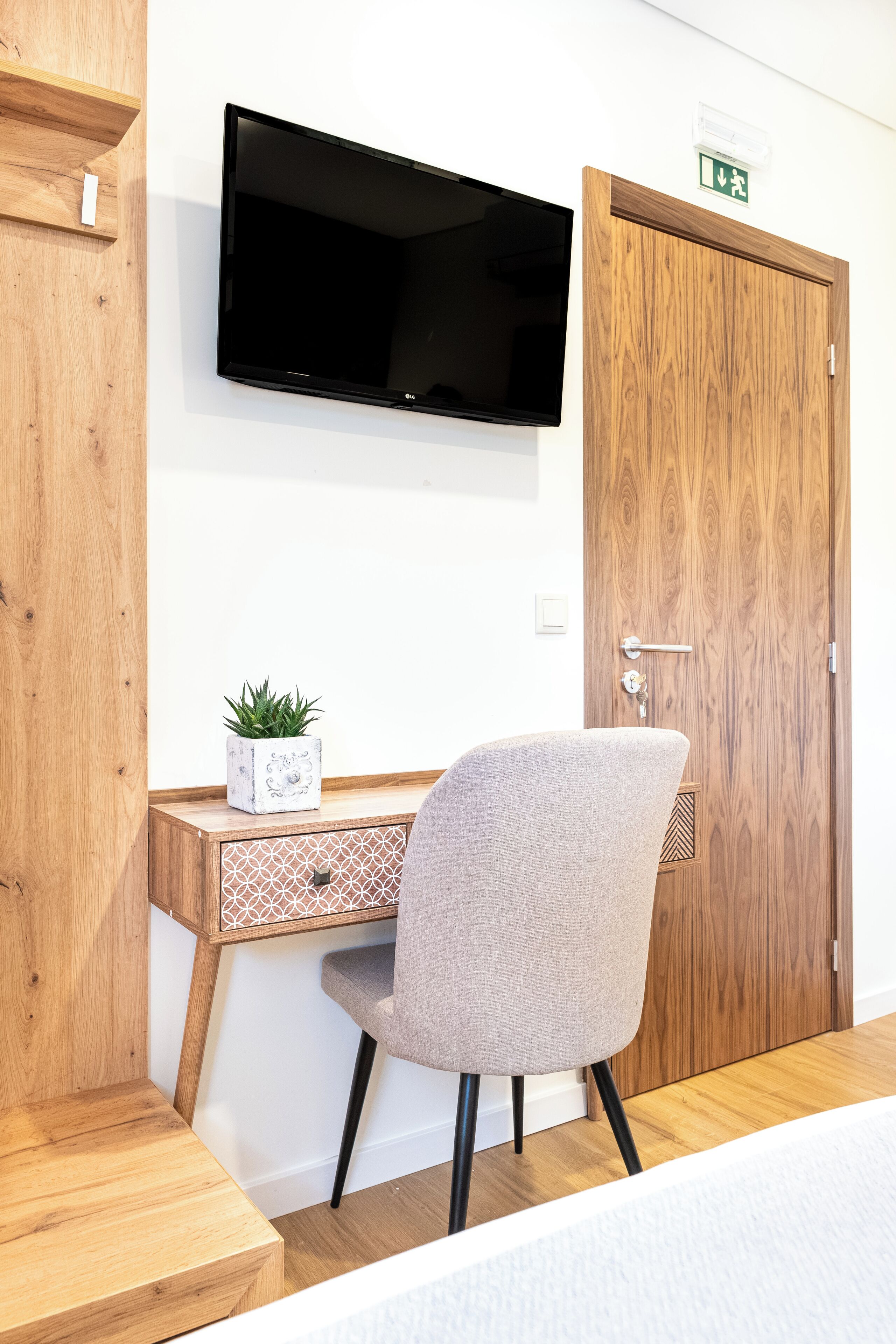 A room with a wooden desk, a grey chair, a flat-screen TV mounted on the wall, and a wooden door with a green exit sign.