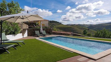 Una piscina rectangular con un muro de piedra y un césped con sillones de descanso y una sombrilla.