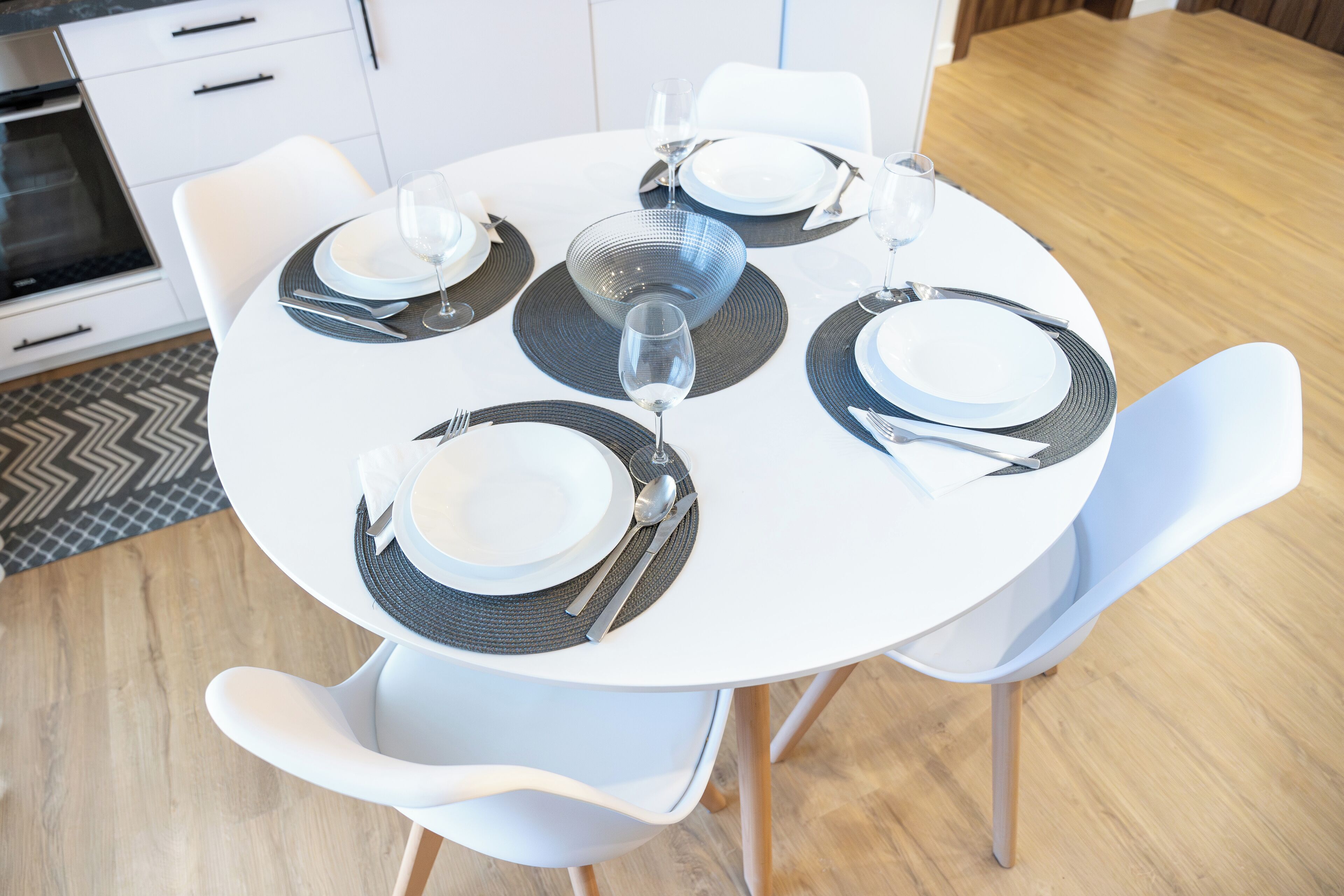 A round dining table with white chairs, set with plates, cutlery, and wine glasses.