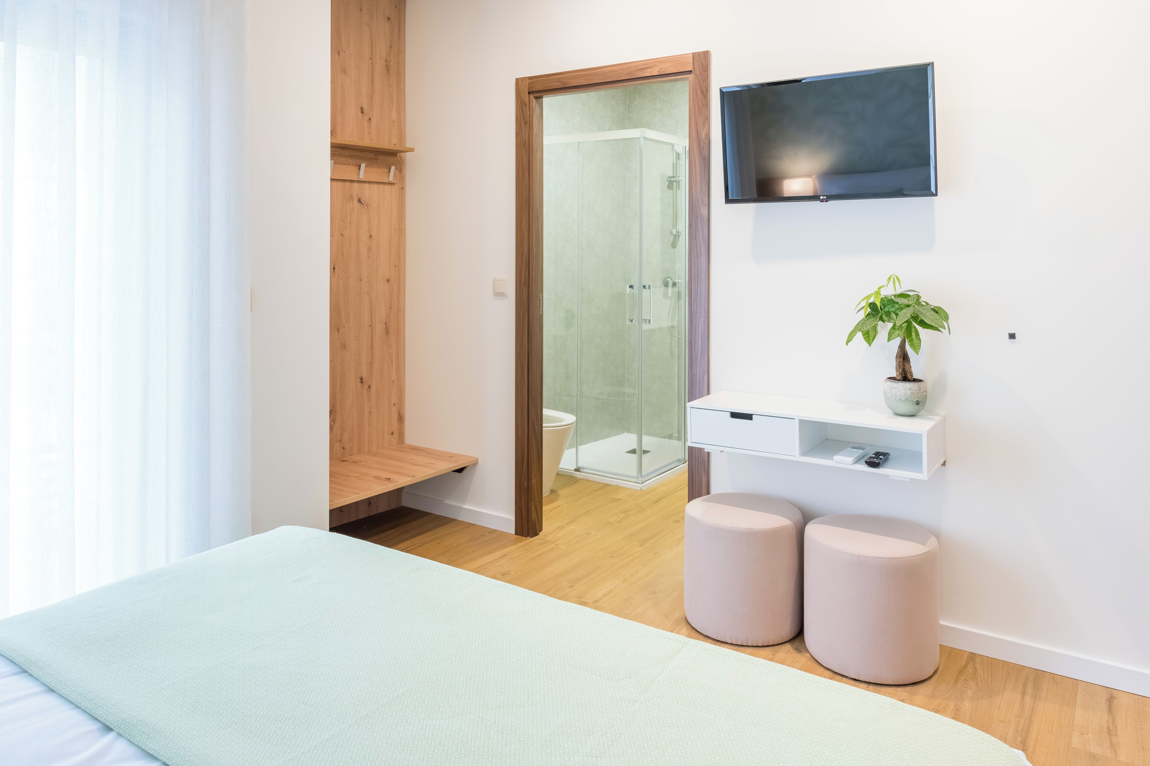 A modern bedroom with a bed, a wooden wardrobe, a glass shower enclosure, a mounted TV, and a small plant on a shelf.
