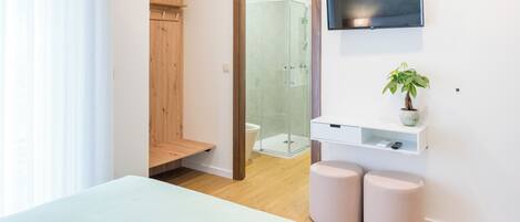 A modern bedroom with a bed, a wooden wardrobe, a glass shower enclosure, a mounted TV, and a small plant on a shelf.