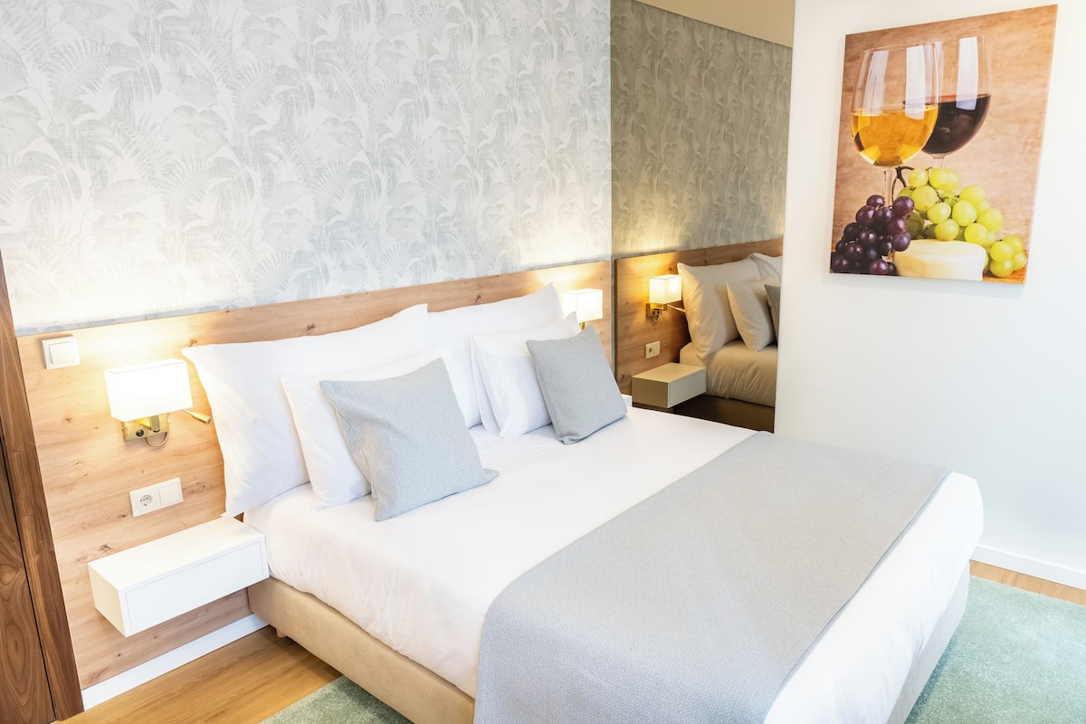 Una cama bien hecha con sábanas y almohadas blancas, cabecera de madera, lámparas de noche y un cuadro en la pared de copas de vino y uvas.