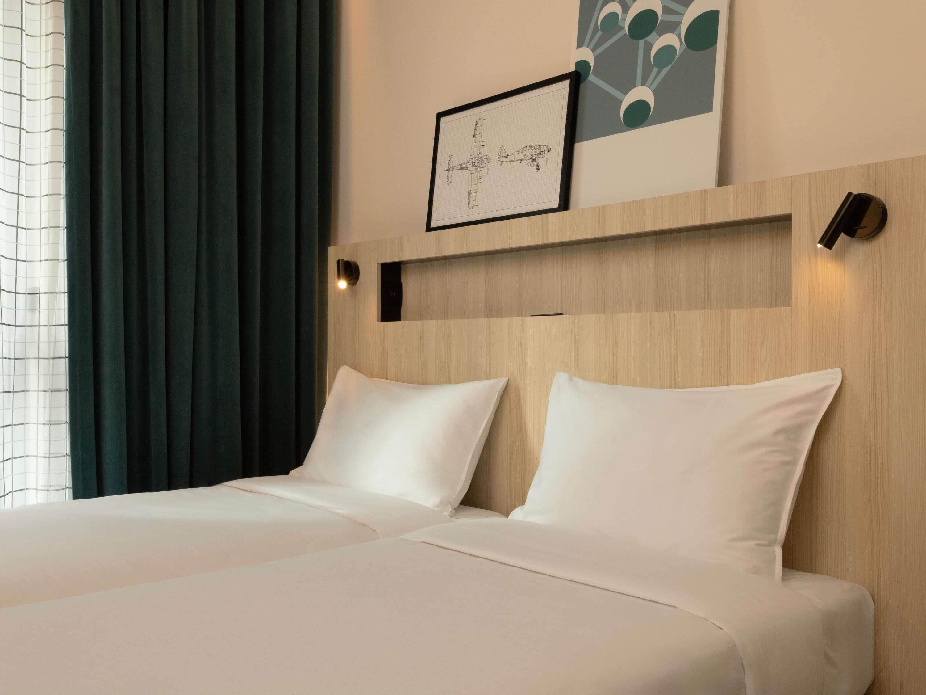 A hotel room with two beds, a large window with curtains, a framed picture on the wall, and a wall-mounted light fixture.